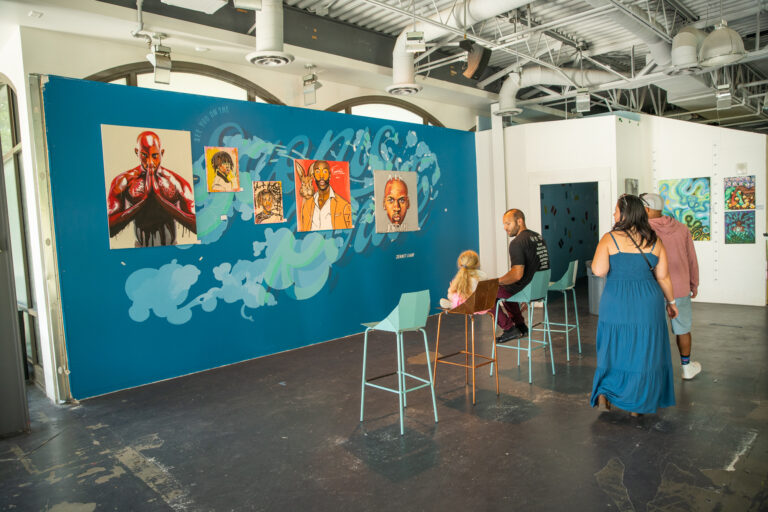 People sitting down viewing paintings hung on a wall that were painted by local Black artists.